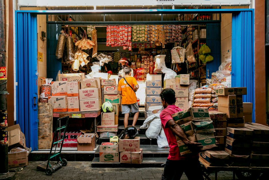 The image depicts the front of a small shop or storage area with a blue metal rolling shutter partially open, revealing a cluttered interior filled with various boxes, bags, and packages stacked on shelves and on the ground. In the foreground, a man in a red shirt is carrying multiple cardboard boxes, while another person in an orange jacket stands near the entrance, facing away and interacting with items or a worker inside. The interior is illuminated with natural daylight, highlighting the assortment of plastic bags, paper packaging, and containers visibly hanging or placed on shelves. The scene appears to be in an urban setting, with the shop’s small steps leading from the narrow street or sidewalk, indicative of a typical market or merchant location. This visual context aligns with private waste handling or rubbish clearance activities, representing the types of clutter managed by services like those offered by Rubbish Removal Lambeth in independent rubbish disposal or clearance assistance for traders and businesses.