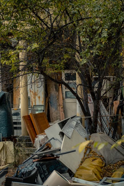A cluttered outdoor scene showing a pile of discarded household waste and debris beneath a tree with green leaves. The pile includes broken wooden panels with warm brown finishes, disused white appliances, and various plastic bags, some yellow, containing unspecified rubbish. A grey, fragmentary piece of fabric or insulation material is draped over one of the appliances. The background features a weathered wooden structure with peeling paint and rusted metal surfaces, indicating a zone awaiting clearance or disposal. The debris is situated on a paved surface, adjacent to a narrow alley or courtyard, surrounded by leaf-covered ground and tangled branches overhead. The scene illustrates an area possibly prepared for a private rubbish removal service like Rubbish Removal Lambeth, highlighting the need for efficient rubbish clearance specifically related to domestic or garden waste on a site requiring on-site or alternative waste handling practices.