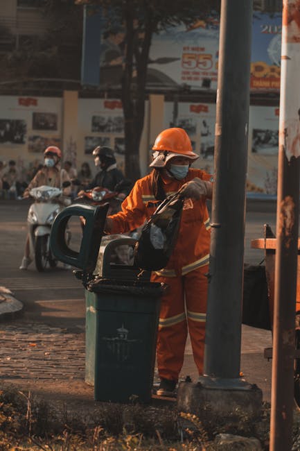 A municipal waste collection worker dressed in an orange high-visibility uniform and a protective helmet, wearing a face mask, is seen standing next to a large green waste bin in an outdoor urban setting. The worker is lifting or placing rubbish into the bin, which is positioned on a paved area near a metal pole and a street curb. In the background, there are several motorcyclists, some of whom are also wearing face masks, riding along a busy road. Behind the road, a billboard with advertisements and images is partially visible, as are trees and a building facade. The scene is illuminated by natural daylight, casting soft shadows and highlighting the textures of the worker’s protective gear and the bin’s smooth plastic surface. This image represents the routine process of on-site waste collection and disposal, illustrating an alternative waste management method often employed by private rubbish removal services like Rubbish Removal Lambeth, in contrast to local authority collection, with the focus on efficient rubbish removal from urban streets.