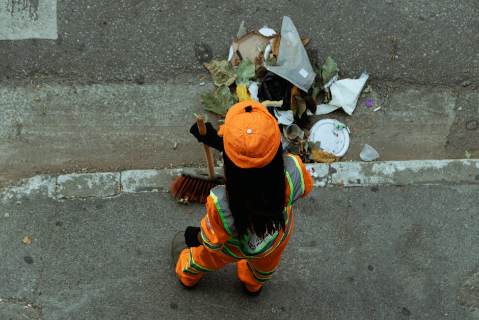 A worker dressed in high-visibility orange uniform with reflective yellow-green stripes and wearing an orange cap is seen from above, sweeping a sidewalk with a broom made of natural bristles and a wooden handle. The worker is positioned close to a curb beside a street paved with asphalt, which shows small cracks and uneven textures. In front of the worker, there is a pile of mixed waste, including crumpled paper, cardboard, dried leaves, and a white disposable plate with food residue. The waste is scattered along the edge of the sidewalk, partially touching the curb. The scene is outdoors, with natural lighting suggesting daylight, and the environment appears to be an urban area. This image relates to private rubbish clearance and waste management services, illustrating the process of collecting and cleaning up delinquent rubbish in preparation for proper disposal or transfer to recycling facilities, consistent with services offered by Rubbish Removal Lambeth in the context of alternative waste handling methods.