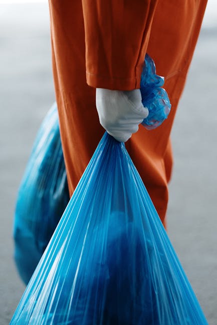 A municipal waste collection worker dressed in an orange high-visibility uniform and a protective helmet, wearing a face mask, is seen standing next to a large green waste bin in an outdoor urban setting. The worker is lifting or placing rubbish into the bin, which is positioned on a paved area near a metal pole and a street curb. In the background, there are several motorcyclists, some of whom are also wearing face masks, riding along a busy road. Behind the road, a billboard with advertisements and images is partially visible, as are trees and a building facade. The scene is illuminated by natural daylight, casting soft shadows and highlighting the textures of the worker’s protective gear and the bin’s smooth plastic surface. This image represents the routine process of on-site waste collection and disposal, illustrating an alternative waste management method often employed by private rubbish removal services like Rubbish Removal Lambeth, in contrast to local authority collection, with the focus on efficient rubbish removal from urban streets.