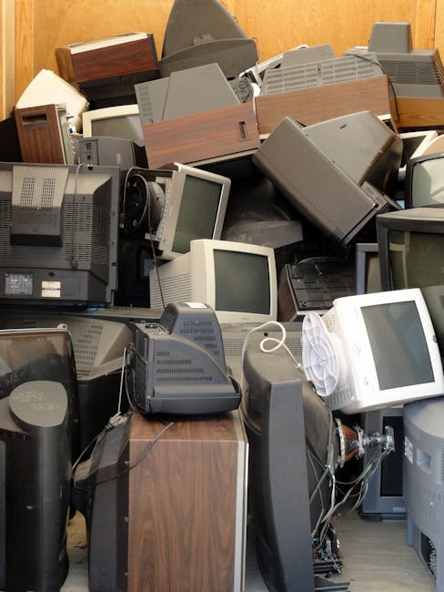 A large pile of discarded electronic waste, including old CRT computer monitors, bulky desktop units, and vintage television sets, stacked haphazardly against a wooden wall. The monitors and TVs display various screens or are turned off, with some showing visible screens and others with plain black or grey surfaces. The electronic devices predominantly feature grey, beige, black, and brown plastic casings, with textures ranging from smooth to slightly textured surfaces. In the foreground, some monitors are partially leaning or stacked against other items, with loose wires and cables tangled among the devices. Several pieces of electronics, including a vintage telephone and other peripherals, are visible within the clutter. The environment appears to be an indoor storage or waste collection area, suitable for handling bulky rubbish like obsolete electrical appliances. The scene depicts the kind of accumulation addressed by private waste removal services such as Rubbish Removal Lambeth, which provides alternative solutions for electronic waste disposal outside standard council collections, emphasizing the importance of responsible handling and clearing of such items.