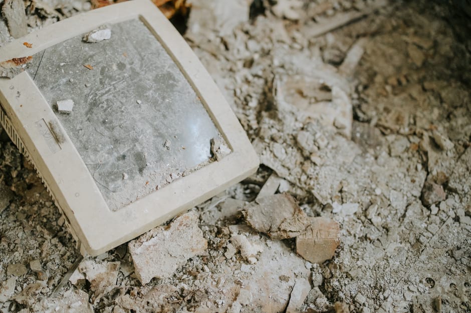 A close-up of an old, discarded computer monitor lying face down on a surface of broken bricks, concrete fragments, and dust. The monitor's beige plastic casing appears worn and dirty, and the glass screen is covered with dust, small debris, and grime, indicating it has been abandoned or left outdoors for some time. Surrounding the monitor are uneven piles of rubble, including chipped bricks, gravel, and dirt, characteristic of a construction site or an area awaiting rubbish removal. The background shows more loose debris and soil, suggesting an environment not maintained for regular waste collection. The scene is illuminated with natural light, highlighting the textures of the dusty, rough materials. This image visually underscores the importance of professional rubbish removal services, such as those provided by Rubbish Removal Lambeth, in handling bulky or hazardous electronic waste as part of an alternative waste management process beyond conventional council collection.