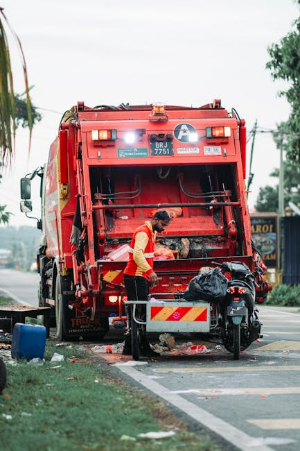 A sanitation worker wearing a red high-visibility vest and black trousers is standing behind a large red rubbish collection vehicle, which is parked on the side of a street. The worker is leaning forward, handling debris or waste materials that are being loaded into the vehicle's rear compartment. The back of the vehicle features reflective yellow and red stripes, headlights, and various identification labels, with the vehicle appearing well-maintained and fitted with waste disposal equipment. Nearby, there is a small black motorbike parked parallel to the vehicle, with a black plastic bag placed on its seat, presumably containing waste or collected rubbish. The surrounding environment includes a paved street with a curb and grassy verge, visible electrical poles and wires, and some trees and buildings in the background, suggesting an urban or suburban setting. Overcast lighting conditions create a neutral, professional atmosphere appropriate for depicting private waste collection or rubbish removal services, consistent with the activities of companies like Rubbish Removal Lambeth engaged in independent collection or alternative waste handling solutions.