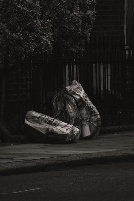 A black and white photograph showing three large garbage bags placed on a paved sidewalk beside a dark iron fence. The bags appear to be made of plastic and are filled with waste, with some displaying printed text or logos on their surfaces. Behind the fence, dense foliage from a leafy tree is partially visible, indicating an outdoor residential or urban setting. The scene suggests a scheduled or on-site rubbish removal process, as might be handled by companies like Rubbish Removal Lambeth, emphasizing the importance of proper waste disposal and alternative rubbish collection methods in the area.