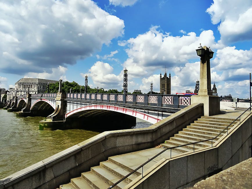 A concrete staircase with metal handrails descending to a riverside walkway, set against a backdrop of the Westminster Bridge spanning the River Thames in London. The bridge features a decorative metal railing painted in light pink with black accents, supported by stone pillars topped with ornamental lamps. The river below displays murky green water with subtle ripples, and the sky overhead is filled with large, fluffy white and grey clouds allowing patches of blue to peek through. In the background, the historic layout includes the Palace of Westminster with its prominent clock tower, part of the London skyline, while the environment suggests a public riverside promenade. The overall scene is illuminated by natural daylight, emphasizing the textures of the stone, metal, and water, and subtly reflects the urban setting where independent waste removal services, such as those offered by Rubbish Removal Lambeth, might operate to support city maintenance and cleanup efforts in busy, historic areas.