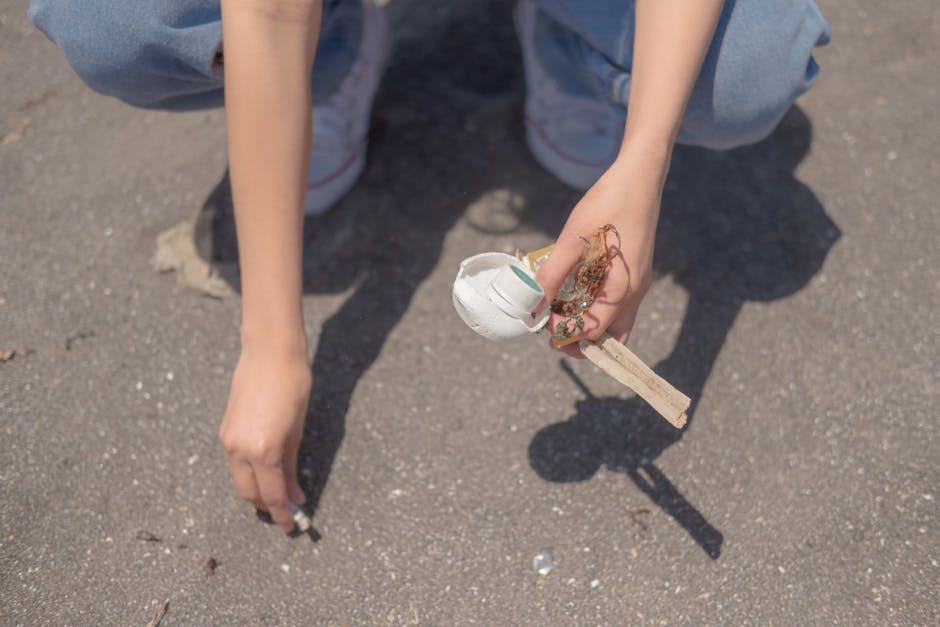 A person wearing light blue denim jeans and white sneakers is crouching on a paved surface, holding a small pile of burnt and partially melted debris consisting of a white ceramic or plaster object, some charred organic material, and a wooden stick. The debris appears to be remnants of a small combustion or fire, with the burnt item showing a smooth, curved surface, and the organic material being blackened and brittle. The wooden stick is light-colored, with a clean cross-section, and is held loosely between the person's fingers. The surrounding environment is an outdoor paved area with a neutral, textured asphalt surface. The scene suggests a context of waste disposal or collection, with the individual possibly preparing waste for collection or removal, connecting naturally to professional rubbish removal services such as those provided by Rubbish Removal Lambeth.