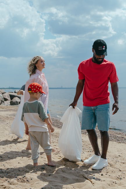 A man wearing a black cap, red t-shirt, denim shorts, and white sneakers is standing on a sandy beach, holding a large white plastic rubbish bag in his right hand. To his left, a young boy dressed in a grey and white outfit with a red cap featuring yellow polka dots is looking at the bag, with another woman standing behind them, dressed in a light pink top and light-colored hair. In the background, the shoreline extends with a line of rocks and the water's edge, under a partly cloudy sky with shades of blue and white. The scene captures a moment of outdoor rubbish collection, reflecting a focus on waste disposal and environmental responsibility, with the beach environment providing a natural setting for local waste handling efforts.