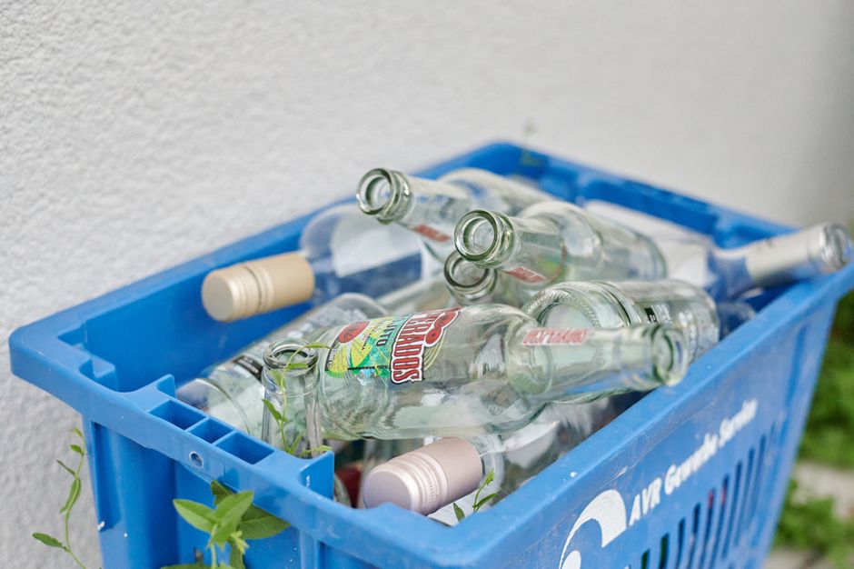 Close-up of a blue plastic rubbish collection bin filled with multiple empty glass bottles, including clear soda bottles with colorful labels and beige plastic caps. The bottles are loosely placed, with some overlapping, and a few small green plant leaves are visible among them. The bin is positioned outdoors against a plain, light-colored wall, and the environment suggests an area for private waste disposal or alternative rubbish collection outside a building. The image emphasizes the handling of recyclable glass bottles as part of waste management services offered by Rubbish Removal Lambeth, suitable for clients considering onsite clearance or independent waste handling options. The scene is well-lit with natural light, highlighting the textures of the smooth glass surfaces and the plastic bin, representative of typical rubbish removal tasks associated with clearing curbside or garden waste.