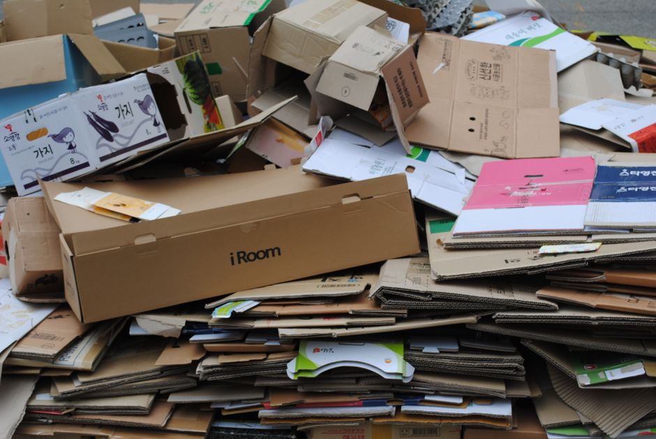 A collection of mixed cardboard boxes and paper-based waste materials stacked behind a blue recycling sign that reads 'RECYCLING ONLY' in white letters, mounted on a light grey shipping container wall. The cardboard includes flattened boxes, partially crushed, with some corrugated edges visible, displaying brown and tan hues with various textures from smooth to rough. Several paper bags and paper packaging are also present, some with printed logos and green accents, slightly crumpled and torn. The waste is arranged in blue and brown recycling bins, with some boxes stacked on top or leaning against each other, indicating on-site disposal or private collection. Overhead, a metal framework partially covers the scene, casting shadows on the waste and container wall. The environment appears to be an outdoor recycling or waste collection area, emphasizing the handling and sorting of waste materials designated for recycling, as would be typical for a rubbish removal service like Rubbish Removal Lambeth.